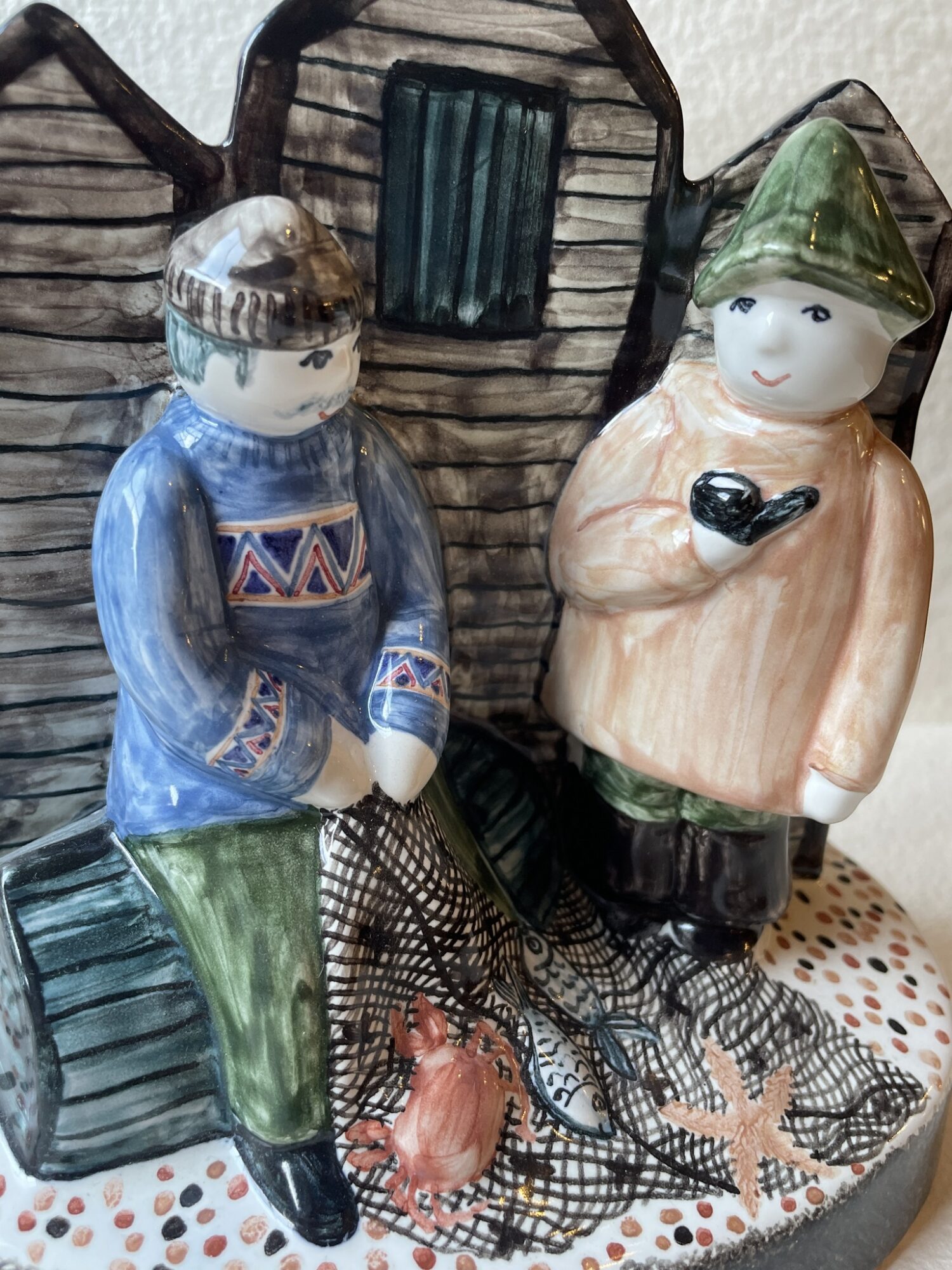 Detail of hand made ceramic figure depicting sussex fisherman mending their nets in front of iconic local black weatherboard fishing huts with fish and shell fish in the nets