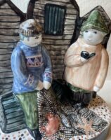 Detail of hand made ceramic figure depicting sussex fisherman mending their nets in front of iconic local black weatherboard fishing huts with fish and shell fish in the nets